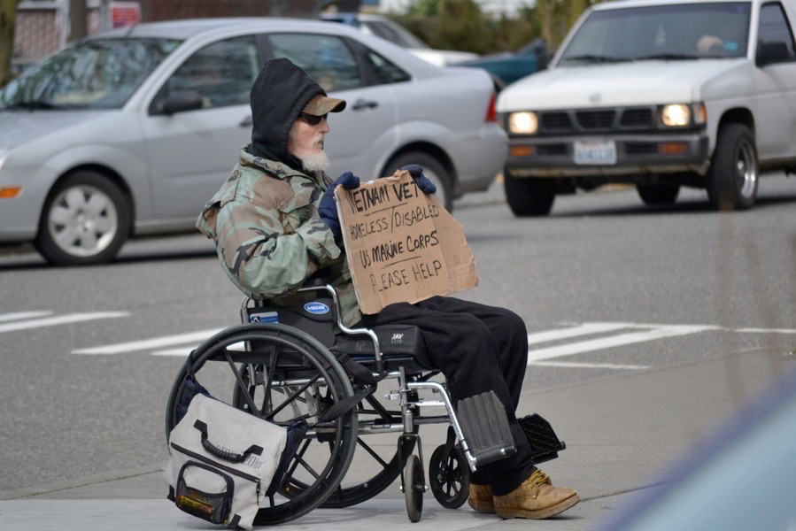 A homeless man in a wheel chair in a crosswalk with a sign that reads ‘Vietnam Vet, homeless, disabled, US Marine Corp, please help“