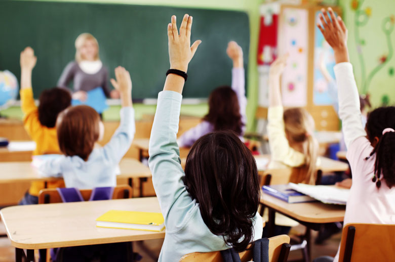 A photograph of children raising their hands in a classroom.