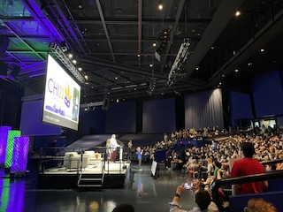 A view of a stage, a large projector screen, and hundreds of people staring at them.