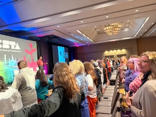 Rows of K-12 CS teachers, arms locked, with a passionate teacher rallying the crowd on stage.