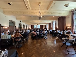 170+ ICER attendees looking toward the end of a dance hall, sitting at round tables.