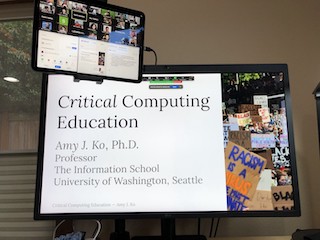 A photograph of a display showing a slide titled "Critical Computing Education", with an iPad hovering above showing Zoom.