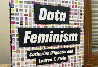 A photograph of the Data Feminism jacket cover, showing the title "Data Feminism", authors Catherine D'Ignazio and Lauren F. Klein, and a backdrop of hundreds of data visualizations.