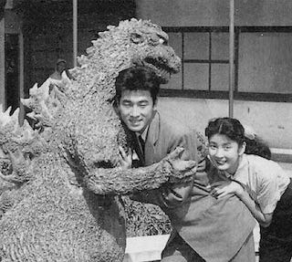 Two Japanese people hugging a human scale godzilla in black and white.