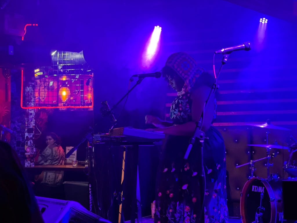 A stage with a Black woman and a keyboard in mics, shrounded in blue.