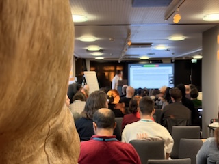 The rear view of the conference room, with 80 heads, partially obscured by the edge of a large wooden stump sculpture.