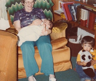 A 1980's photo of a women with curly hair in a recliner nursing a baby with a bottle and a small child holding a stuffed panda on the floor.