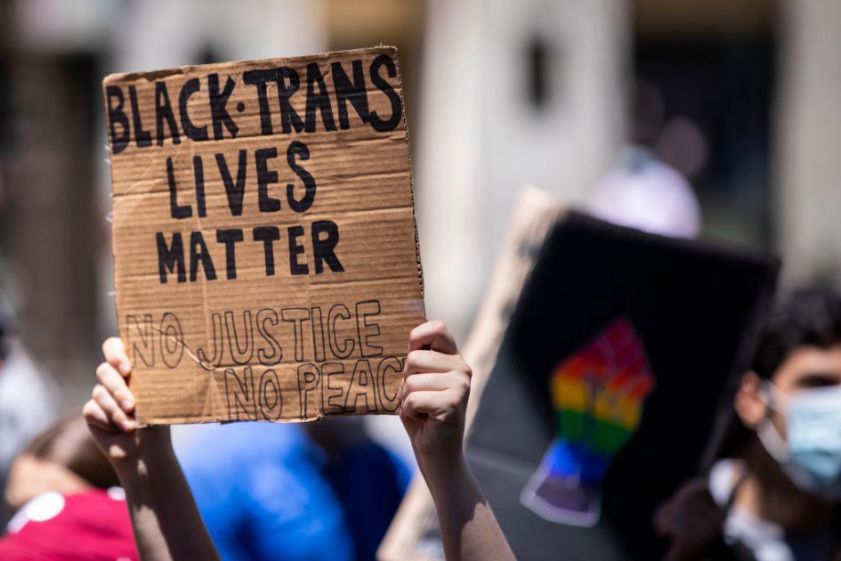A protester holds up their homemade sign on a box that says, 'Black Trans Lives Matter No Justice No Peace'