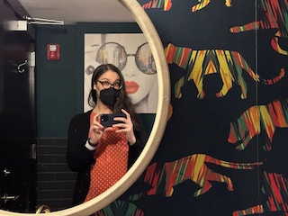 Amy holding her smartphone in front of a round mirror, with a print of a woman in sunglasses with red lipstick and colorful tiger wallpaper.