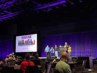 A dark purple stage, a slide that says SIGCSE, and the organizing committee kicking things off.