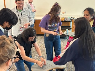 Eight people standing and looking down at a desk with a very small battery powered robot; smiles abound, and a snack cart in the background.